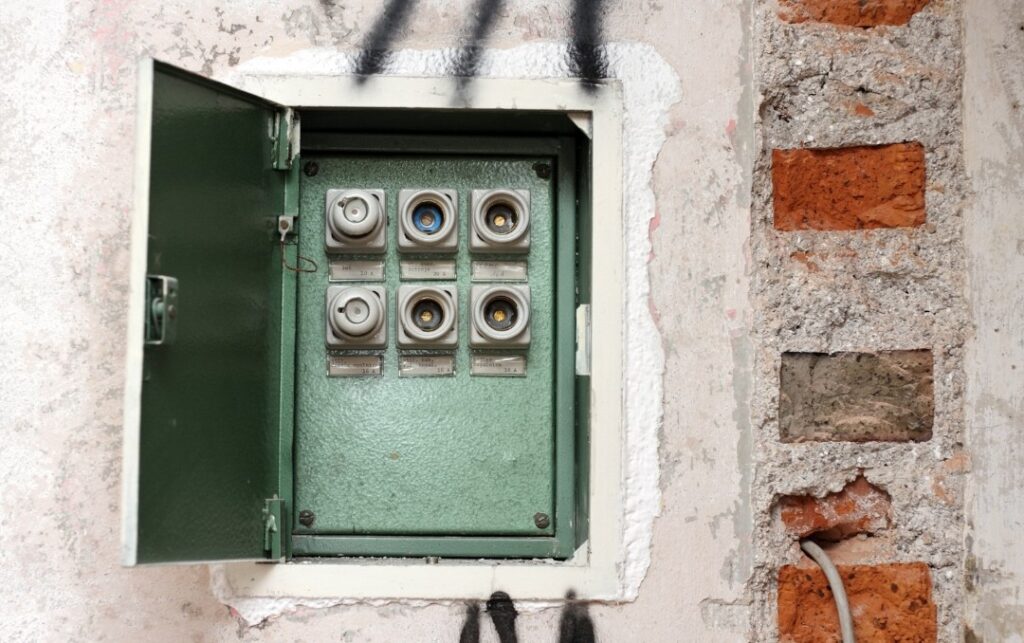 Old fuse box with ceramic screw-in fuses mounted in a green metal panel on a partially plastered brick wall , an example of early 1900s residential electrical systems before circuit breakers.